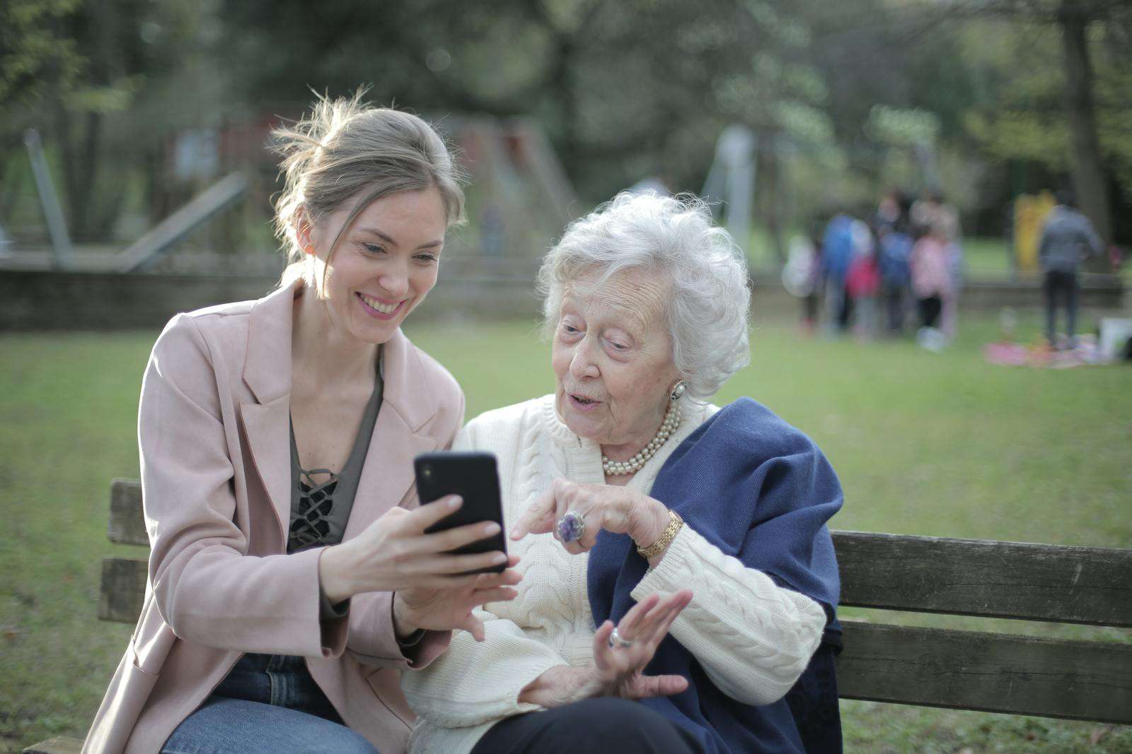 Care jobs Delighted female relatives sitting together on wooden bench in park and browsing mobile phone while learning using