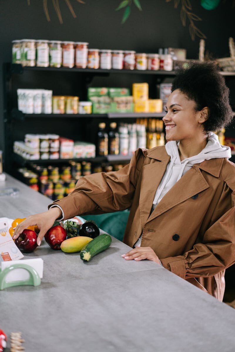 The Beginner's Guide to Reducing Synthetic Additives in Your Daily Diet Woman with afro hair in a brown coat selecting fresh vegetables at a store counter, smiling.