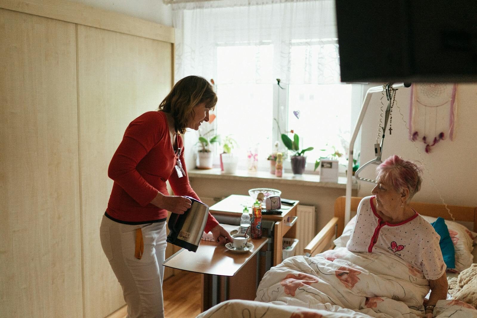 5 Questions To Ask Before Hiring A Live-In Care Provider A caregiver serves tea to a senior woman in a cozy bedroom setting, fostering comfort and care.