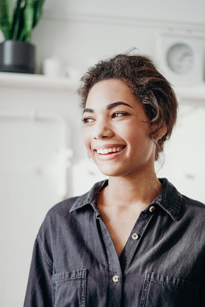The Link Between Oral Health and Overall Health: What the Research Actually Shows A cheerful woman smiling indoors, dressed in casual attire, evoking a sense of happiness and relaxation.