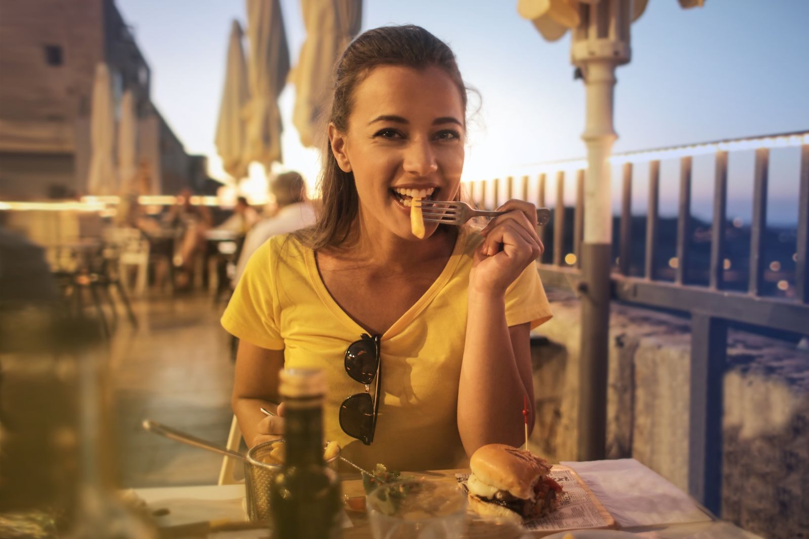 What Makes a City an Inspiring Culinary Destination? Woman in yellow top smiling while dining on a burger at a sunset outdoor cafe.
