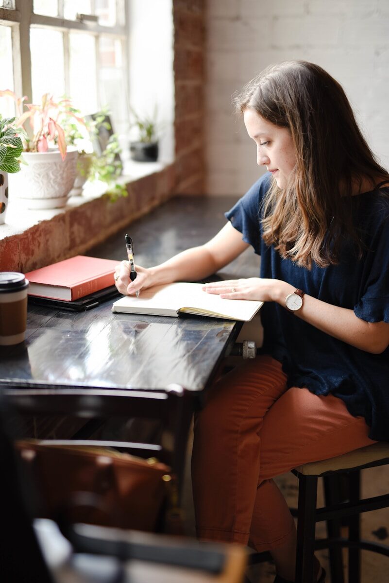 healthy living tips - journaling woman sitting in front of black table writing on white book near window