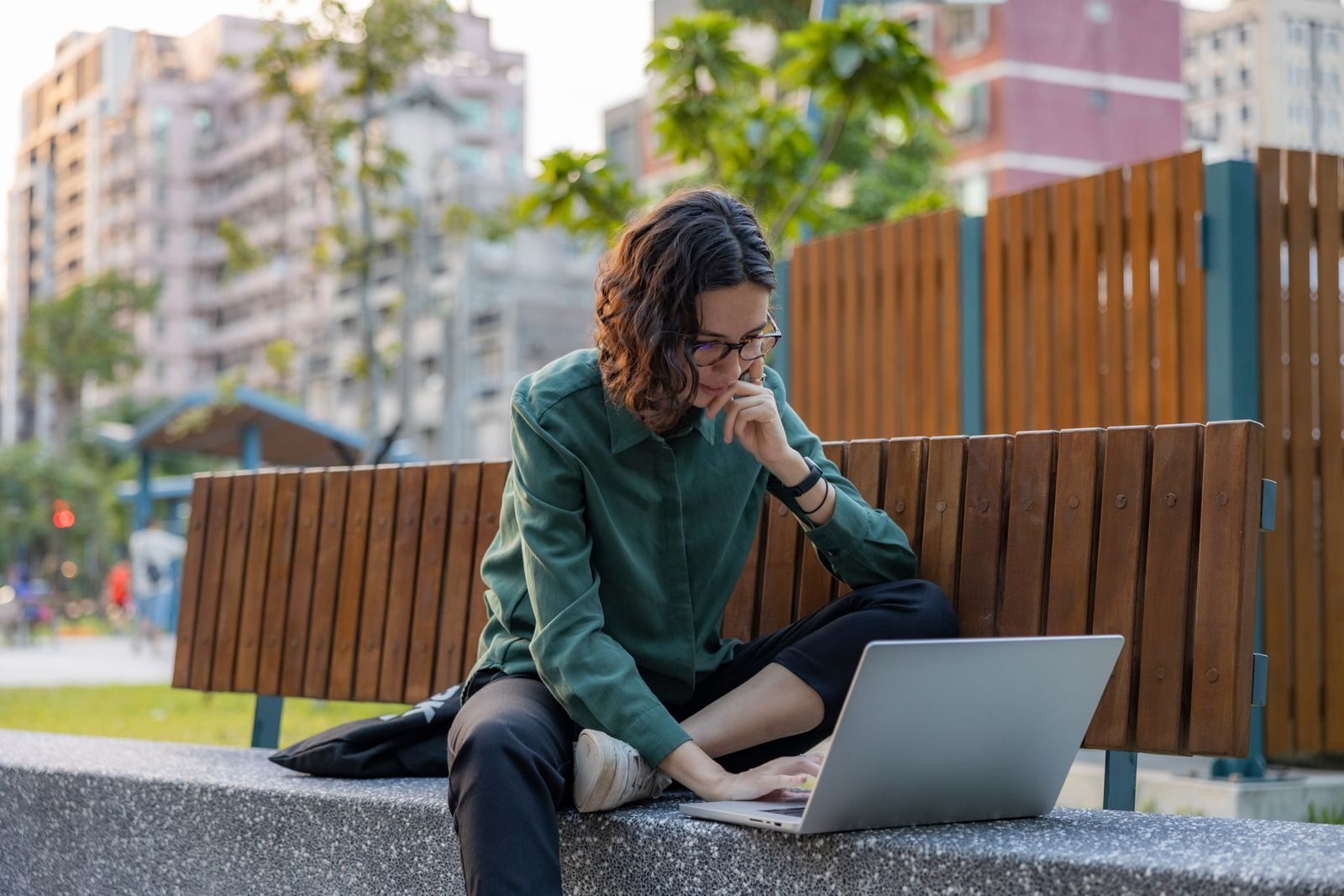 Small Digital Habits That Can Make Everyday Life Easier A young woman works on her laptop while sitting on a bench in an urban park during the day.