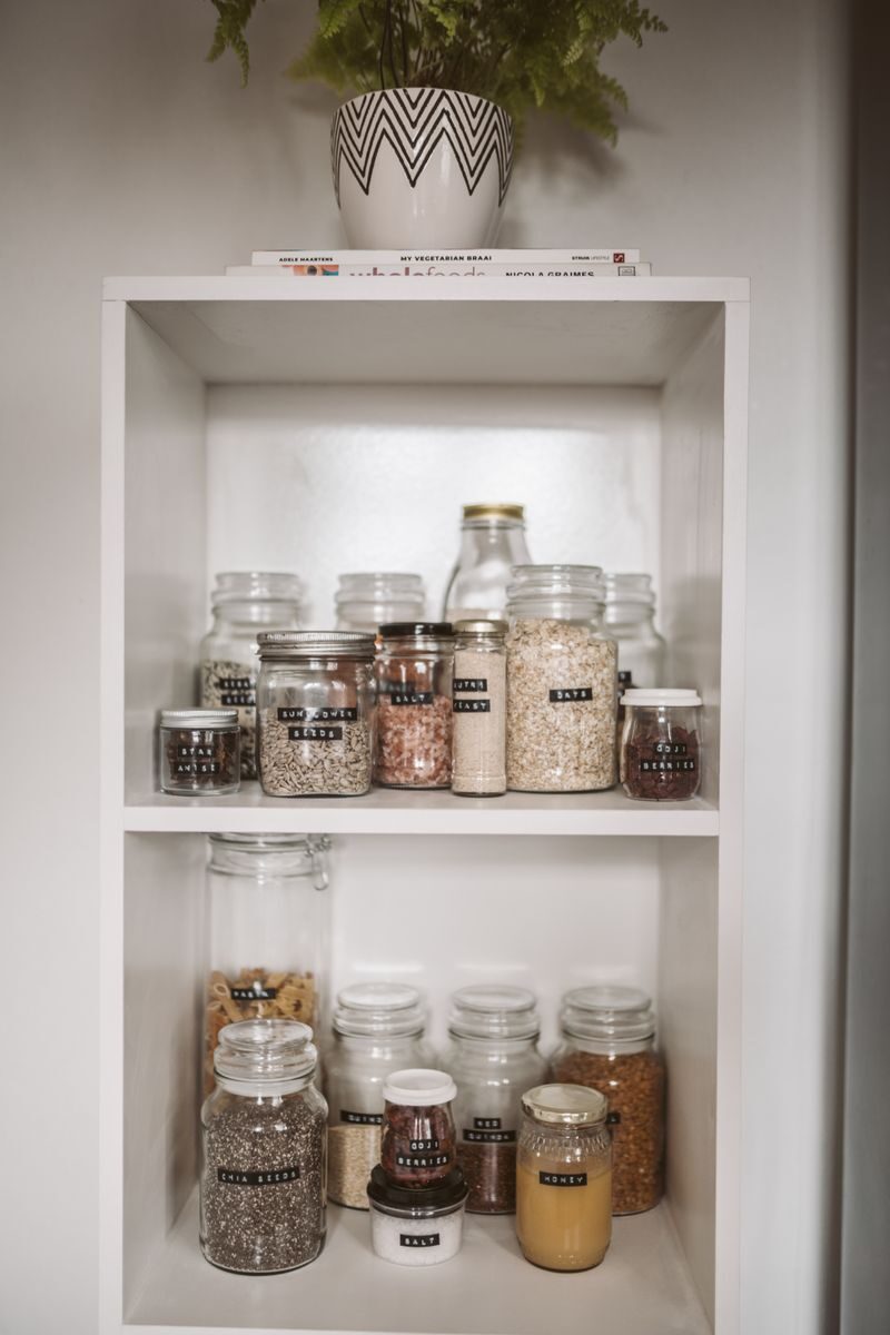 Food Storage Basics For Busy Households Neatly arranged pantry showcasing labeled glass jars with assorted grains and seeds.