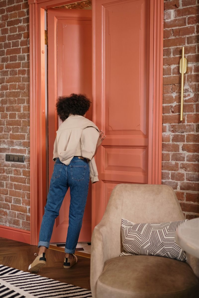 The Calm Home Checklist: Small Repairs That Quiet a Room A woman with curly hair opens a large coral door in a modern loft room with a brick wall.